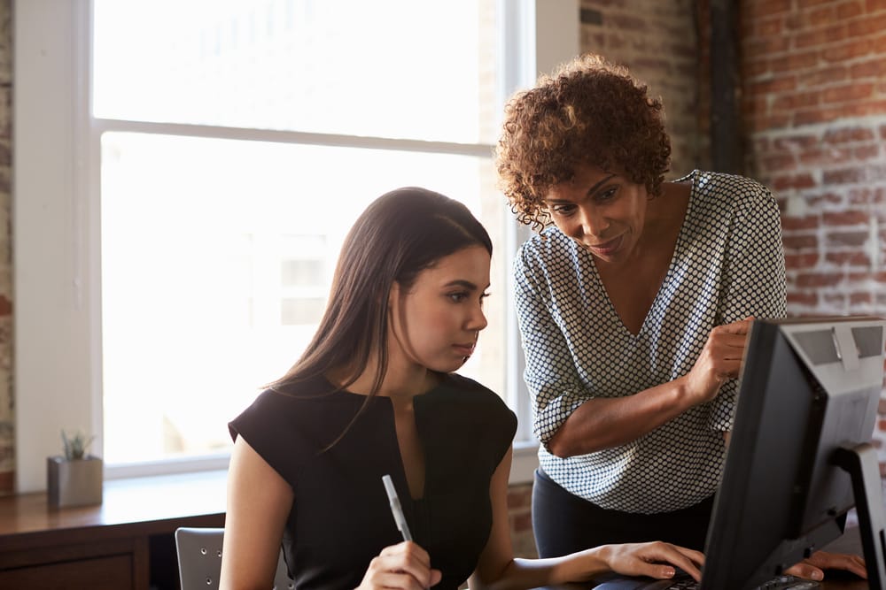 Woman standing looking at a computer screen over the shoulder of another woman, sitting.