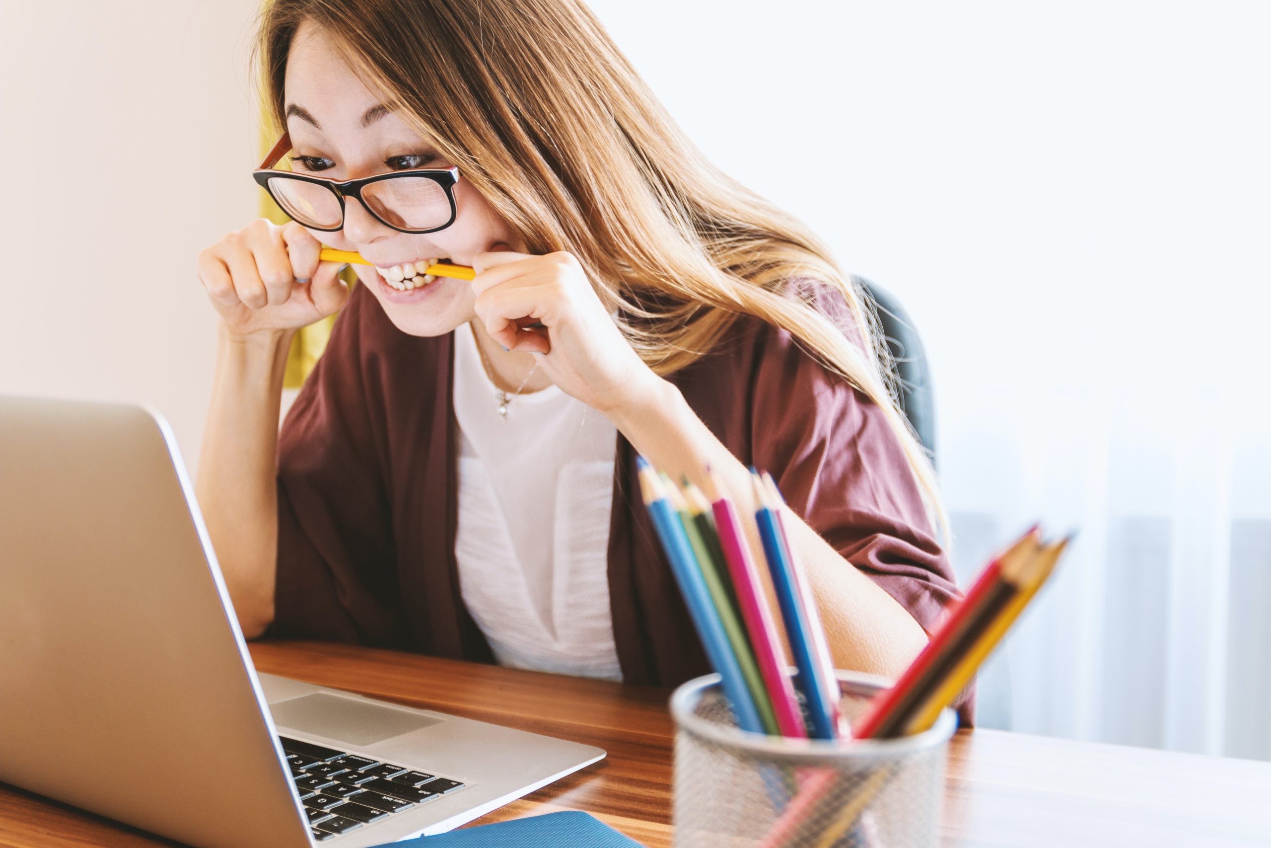 Woman sits at a table nervously biting a pencil whilst looking at a laptop screen. A pot of pencils sits beside the laptop.