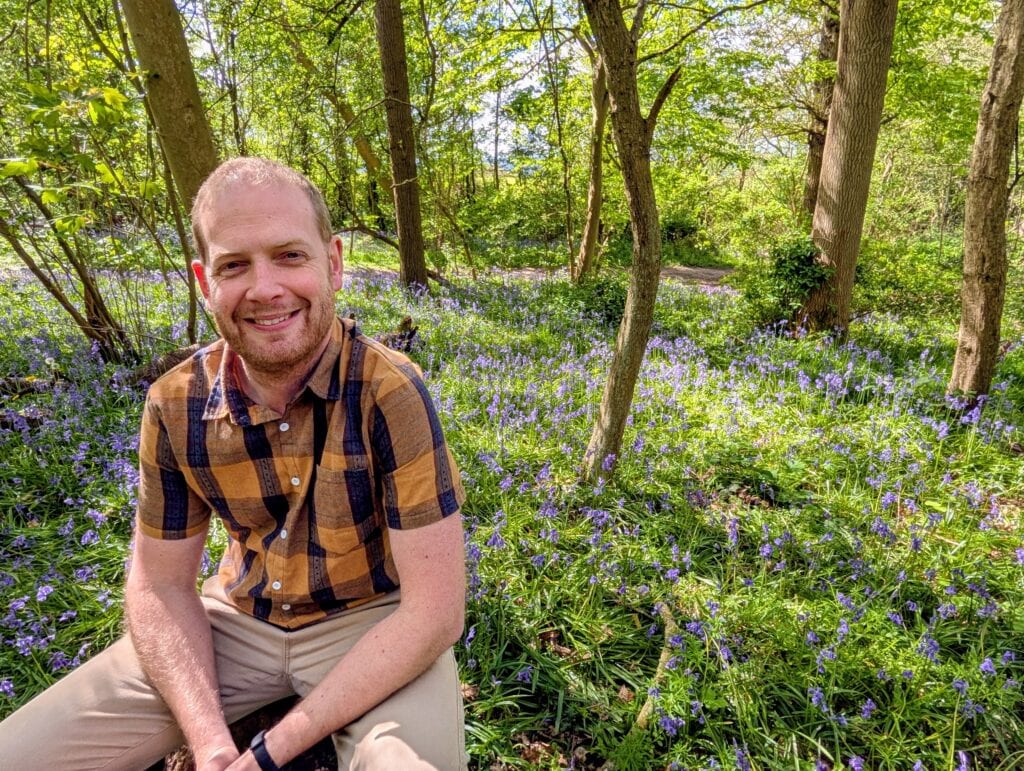 Smling man in a checked shirt with bluebells and trees behind him. CharityJob article on writing the ultimate cover letter.