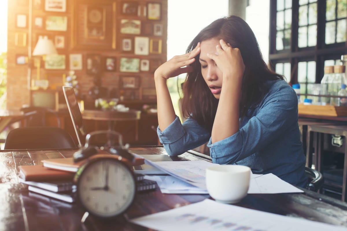 Woman in blue shirt with her head in her hands. There's an alarm clock on the table. 