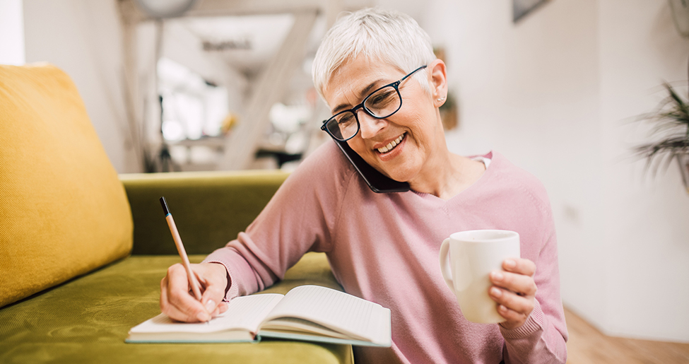 Woman cradling mobile between her shoulder and ear, writing in a notebook, holding a mug