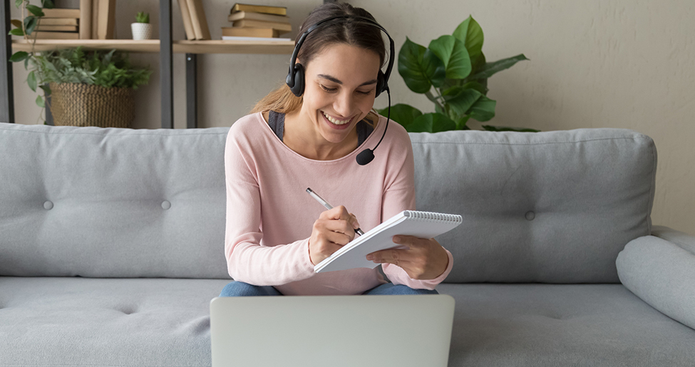 Woman sat on sofa in front of laptop wearing a headset and writing on a pad of paper