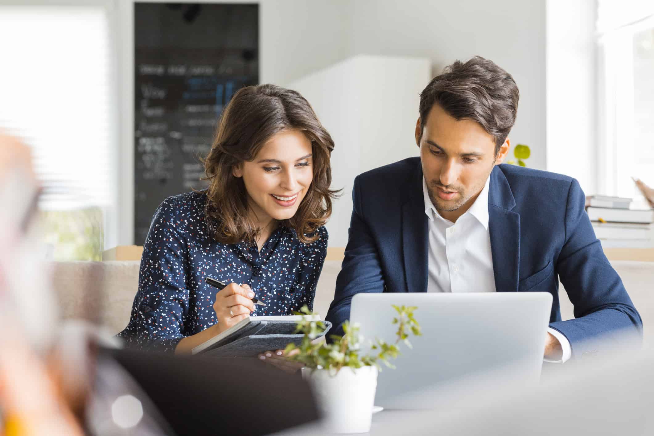 A woman with a notepad and a pen and a man are sat around a laptop looking at the screen