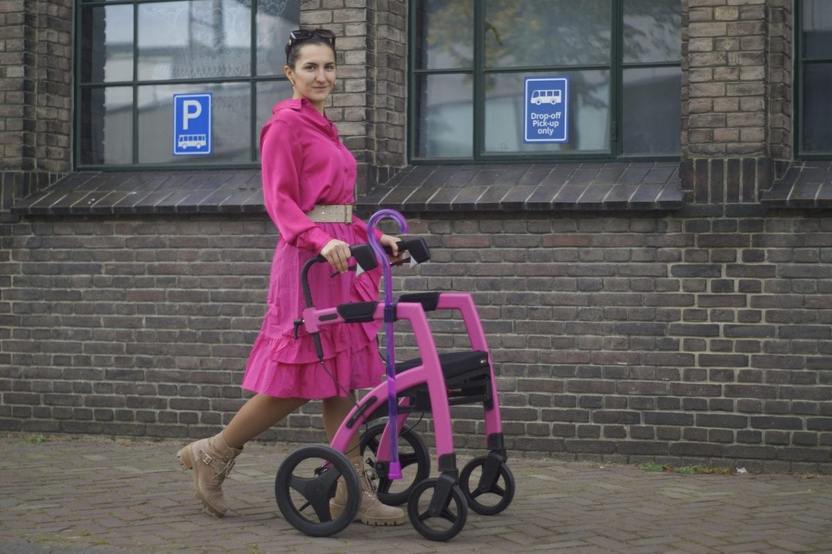 Woman walking outside. She's wearing a bright pink dress and boots, and is using a pink walking aid. CharityJob article on supporting disabled people in the workplace.