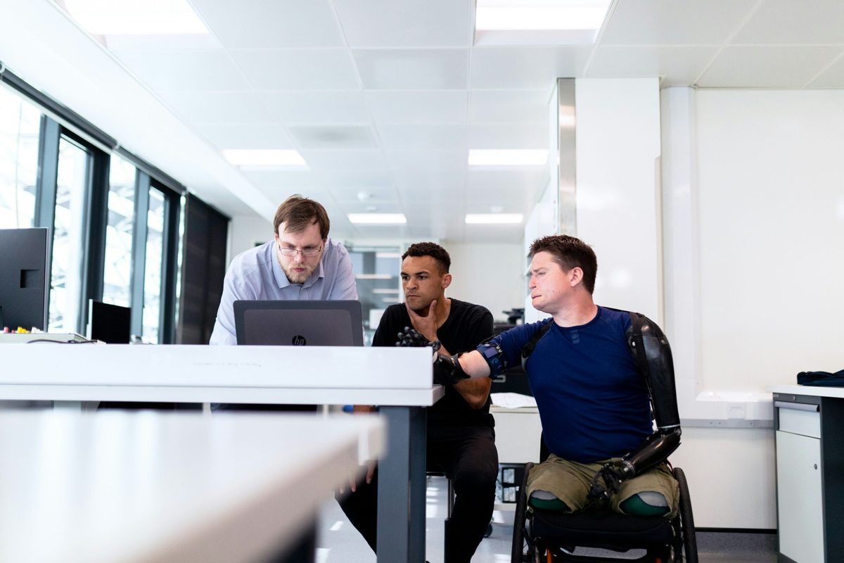 Three men in an office environment looking at a laptop. One of the men is in a wheelchair and has prosthetic limbs. CharityJob article on supporting disabled employees.
