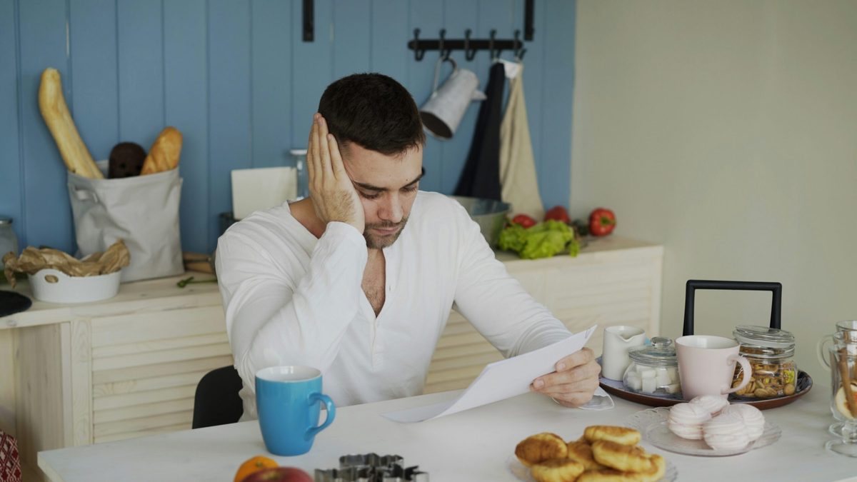 A man in a white top sitting at a table with blue mug and pastries. He's looking at a piece of paper and appears despondent.