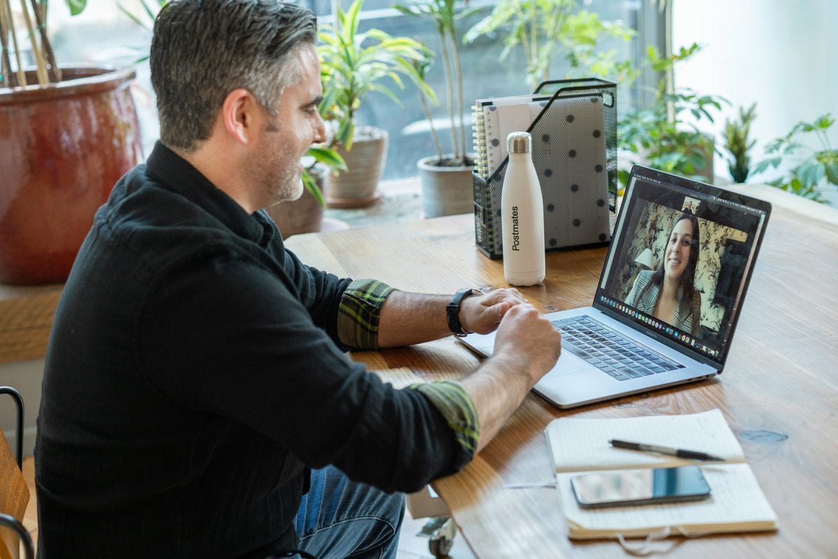 Man in a dark shirt with sleeves rolled up conducting a remote recruitment interview via a laptop. A smiling woman is on the screen of his laptop.
