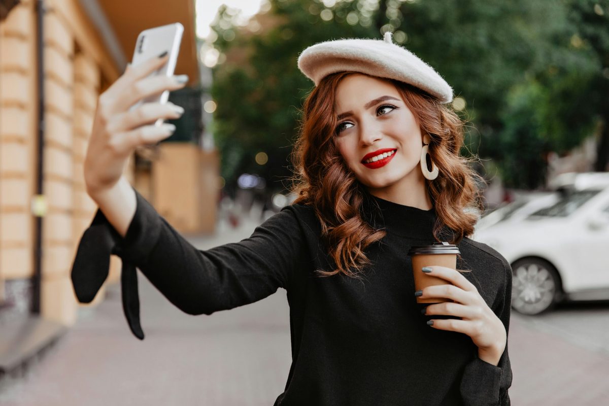 Red headed woman with a beret on her head and a black top. She's holding a coffee and filming herself on a mobile phone. CharityJob article on asking candidates to film video applications.