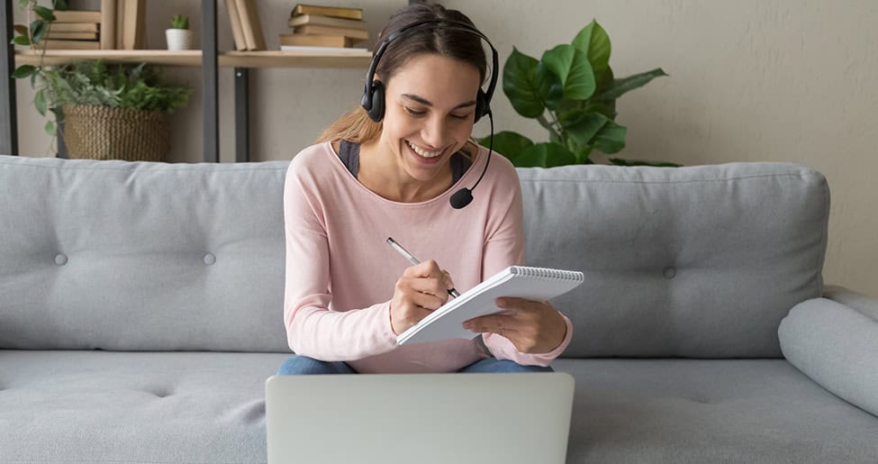 Woman in long-sleeved pink top with headset. She's holding a pad and smiling at a laptop as she does a remote recruitment interview.