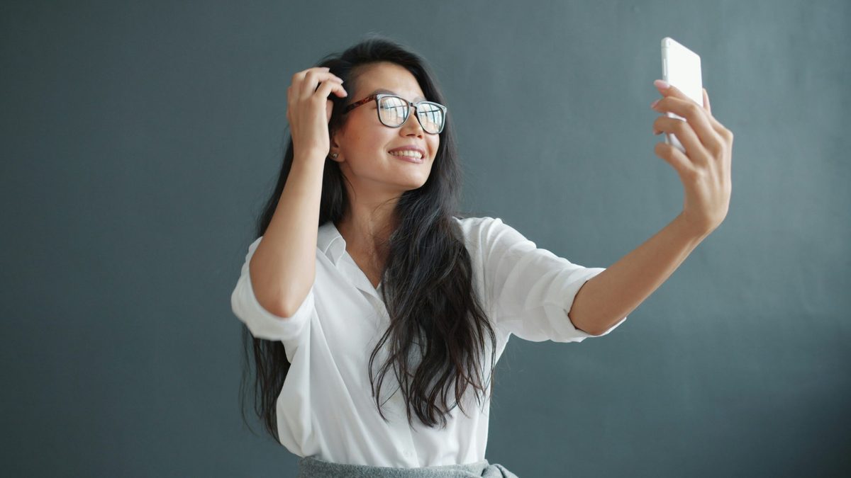 Woman in glasses and white shirt with long dark hair. She's filming herself on a mobile phone