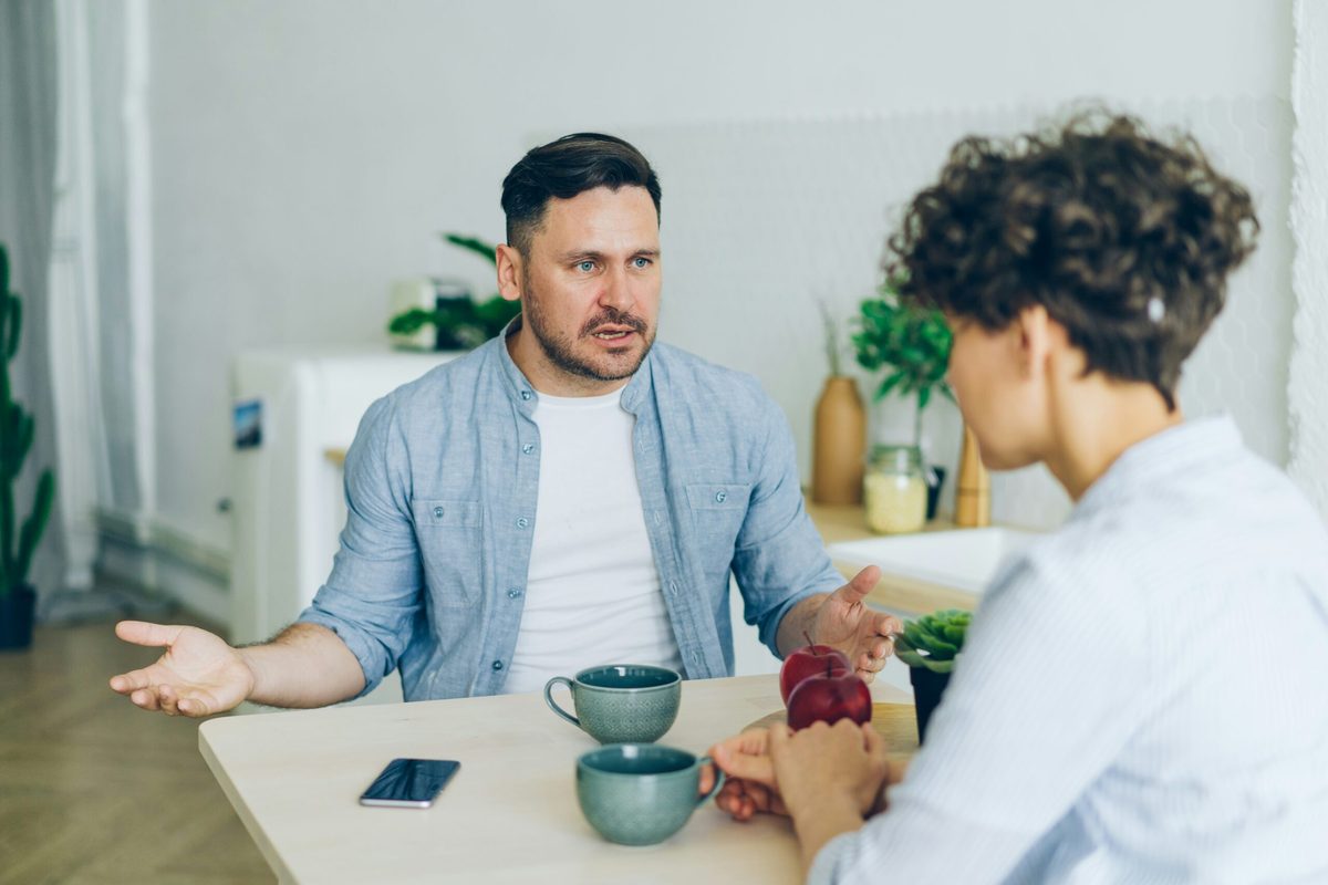 Man gesturing with open arms. He's sitting opposite a woman, and they both have coffee cups. Article on the benefits of mental health first aiders in the workplace. 